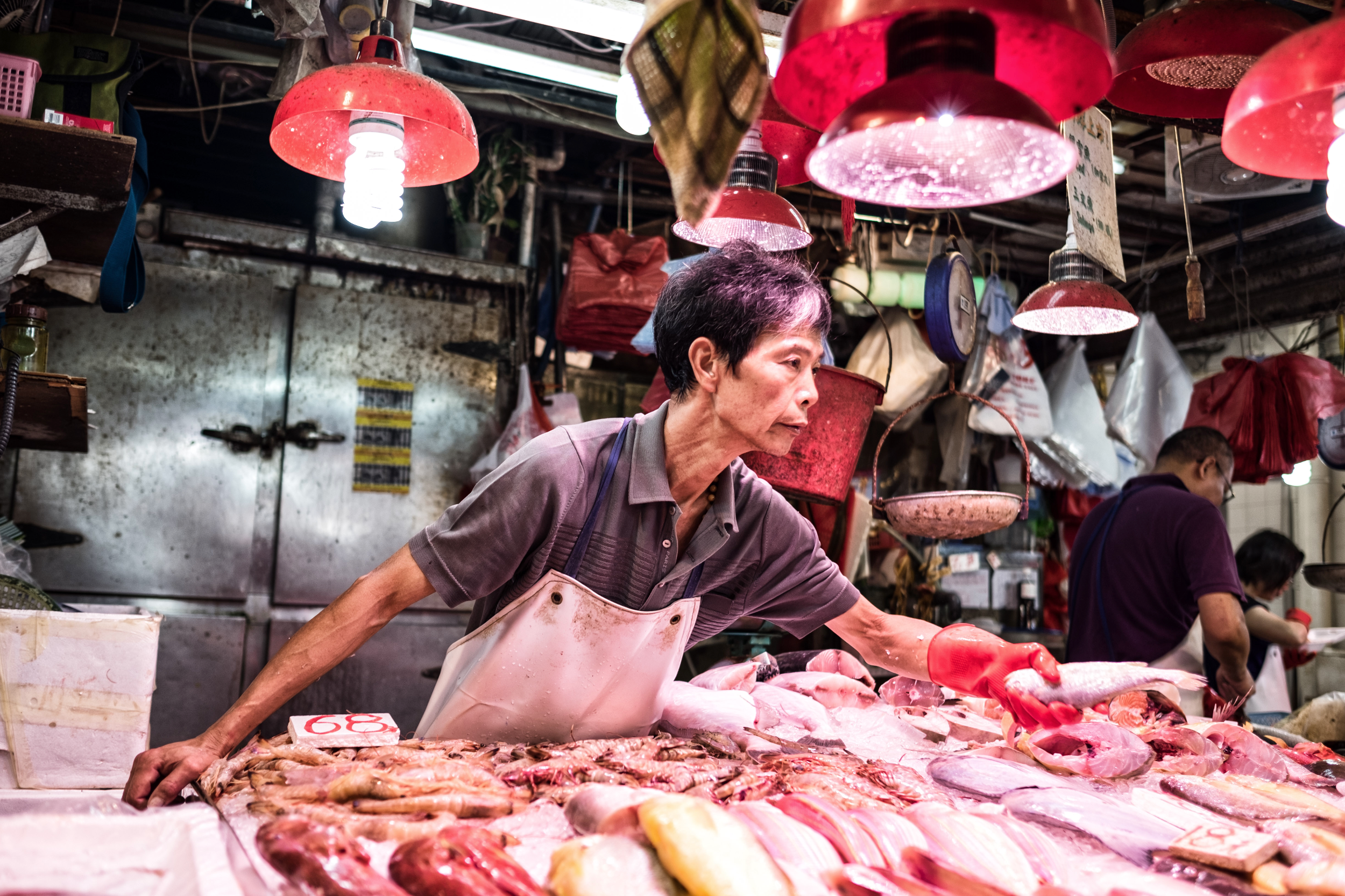 A fish vendor at a wet market reaches across a counter of fresh seafood under red heat lamps, selecting a fish while other vendors work in the background.