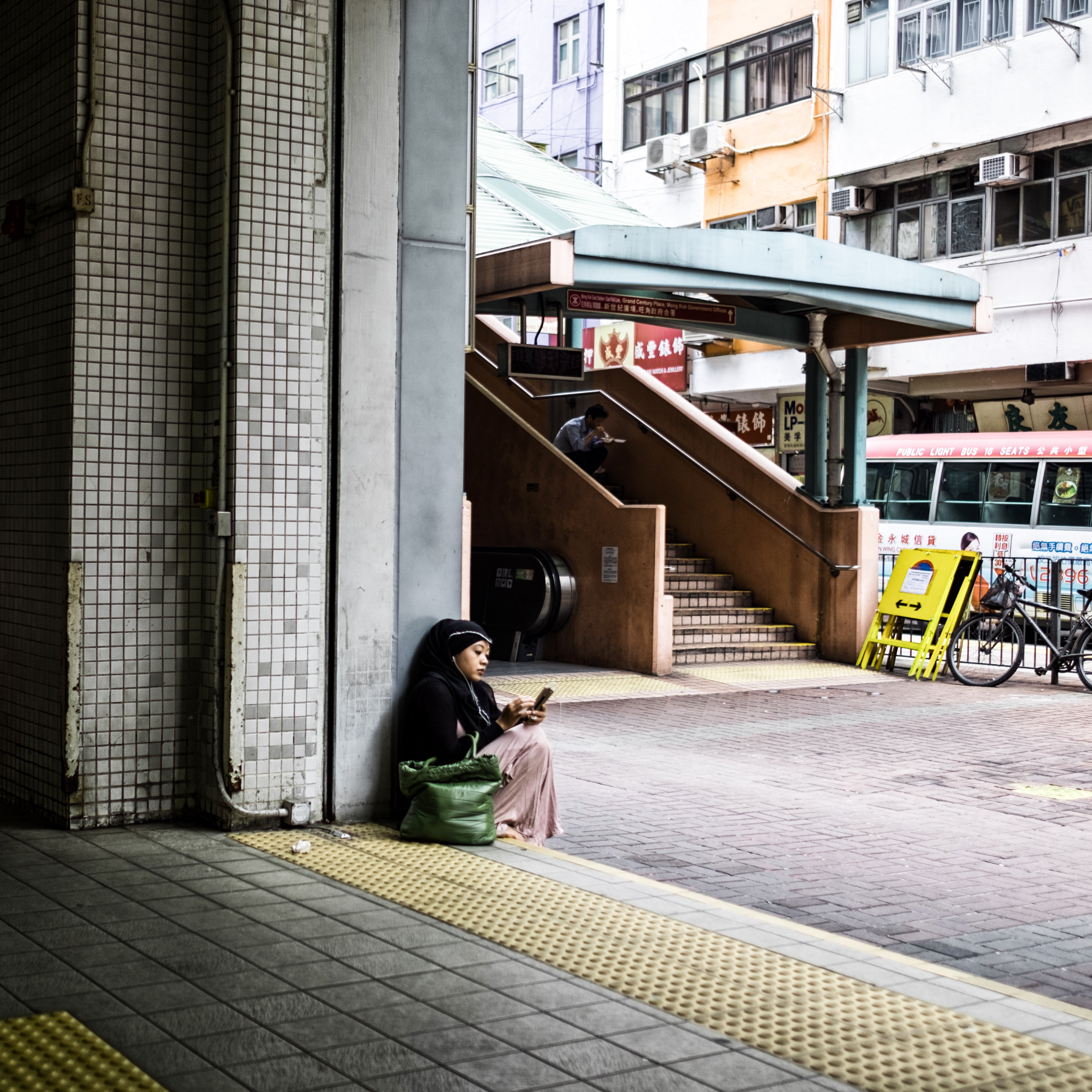 A woman sits on the ground beneath a covered stairway, leaning against a tiled wall while looking at her phone, with city buildings and a bus stop in the background.