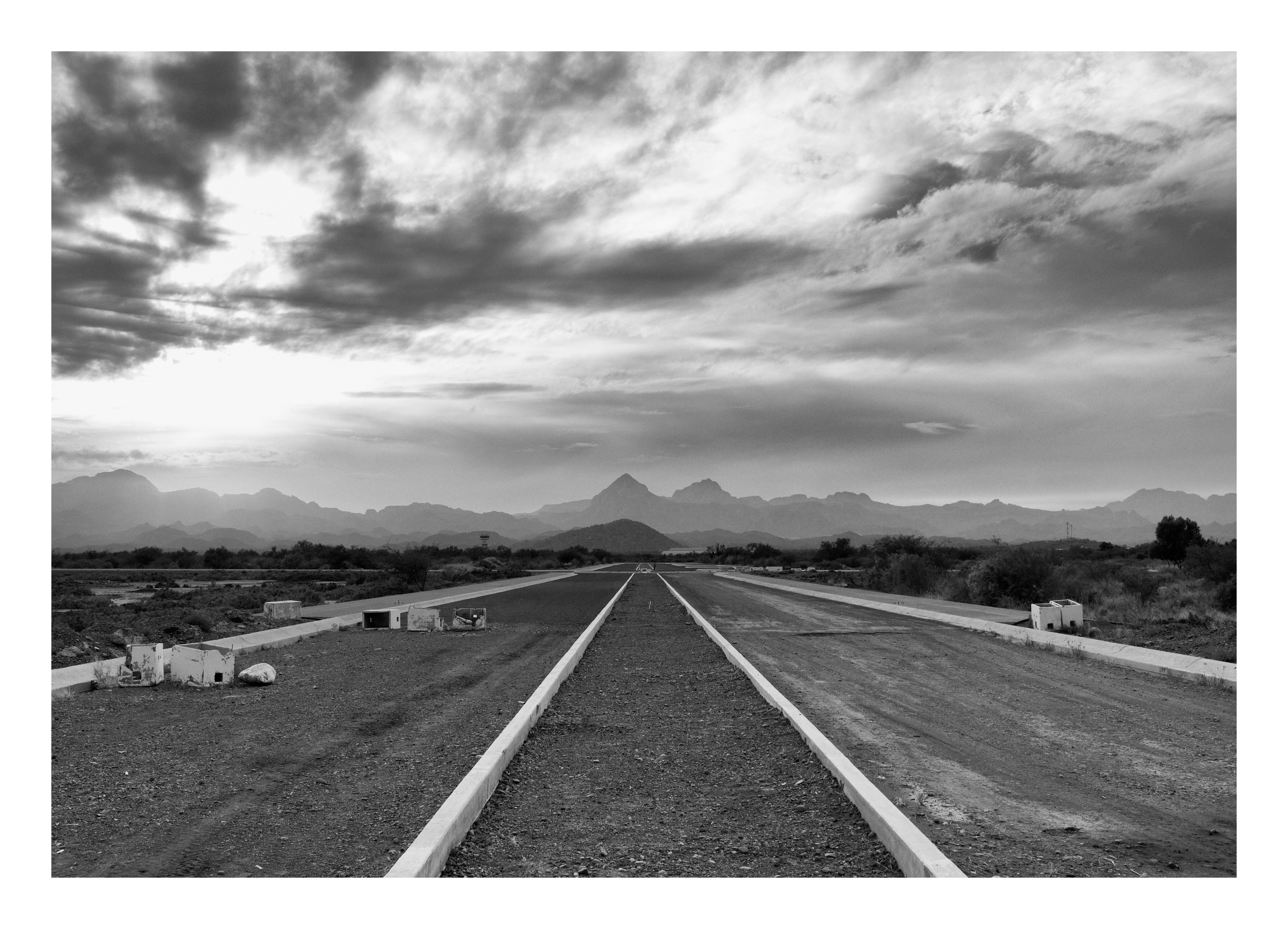 Black and white photograph of an empty roadway or runway stretching toward distant mountains beneath dramatic, layered clouds.