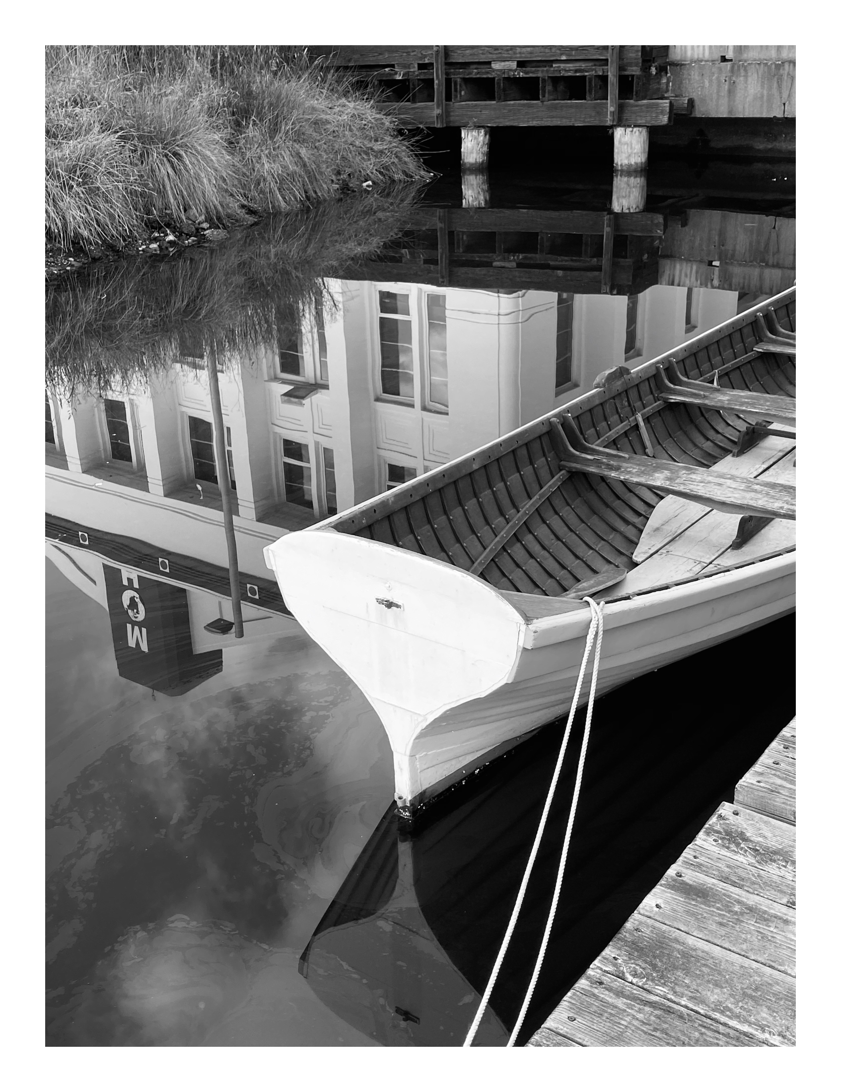 Black and white photograph of a small boat moored beside a wooden dock, reflected in still water alongside nearby buildings.